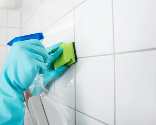 Manos con guantes azules rociando limpiador y frotando juntas de azulejos blancos con una esponja verde para blanquear juntas azulejos.