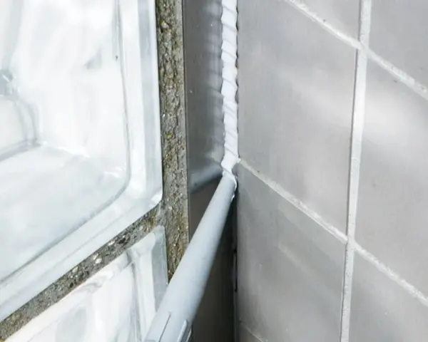 En fogpistol applicerar ny silikonfog längs en vägg av grå kakelplattor och glasblock.
