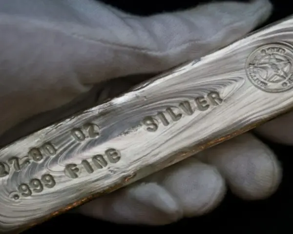 Mano guantata tiene un lingotto di **titolo argento** 999.9, con incisioni che indicano 27.00 oz e "FINE SILVER".