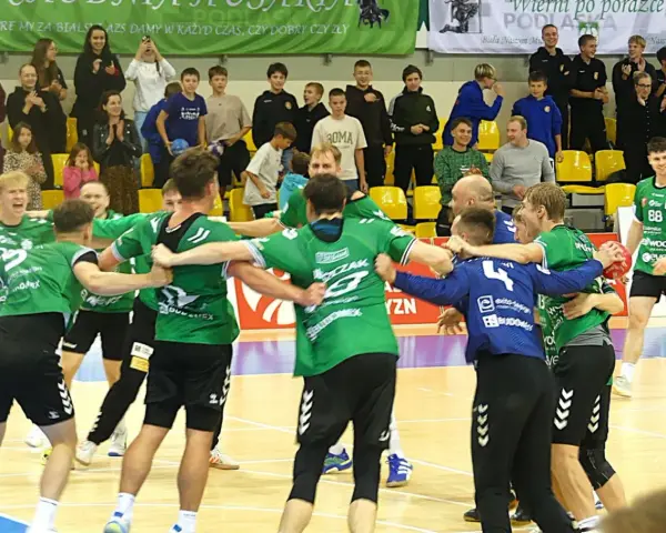 Krystian Woźniak i jego drużyna świętują zwycięstwo. Zawodnicy w zielonych strojach przytulają się na boisku, a na trybunach widać kibiców.