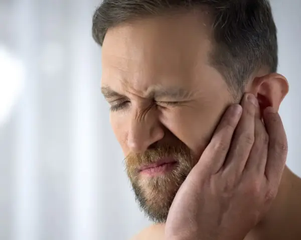 Mężczyzna z zarostem cierpi z powodu bólu chrząstki ucha przy dotyku, zaciskając oczy z bólu.