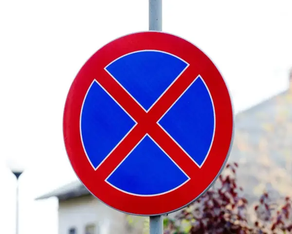 Ostrzeżenie dla kierowców: zakaz parkowania. Znak drogowy w kształcie koła z czerwonym obramowaniem i niebieskim tłem, na którym widnieje czerwony krzyż.