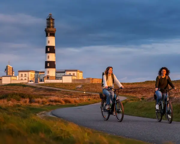 Deux femmes font du vélo sur une route sinueuse à Ouessant, avec un phare emblématique en arrière-plan. Une belle façon de visiter Ouessant à pied, ou plutôt à vélo !