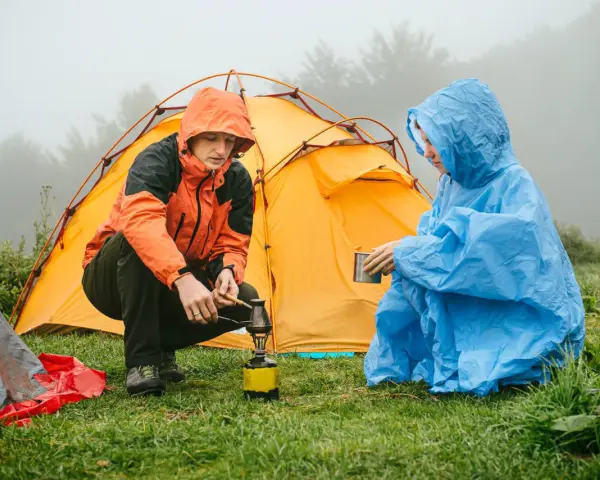 Zwei Camper in Regenponchos bereiten sich auf das Zelten bei Regen vor. Einer kocht auf einem Campingkocher, der andere hält eine Tasse.