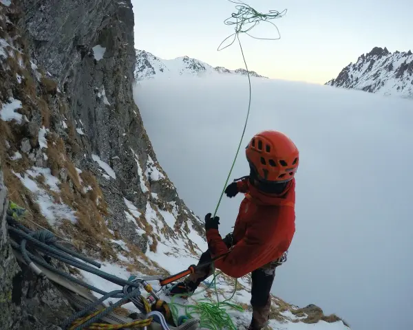 Wspinaczka górska w chmurach. Alpinista w pomarańczowym kasku i kurtce przygotowuje się do zjazdu na linie, otoczony zimowym krajobrazem i mgłą.