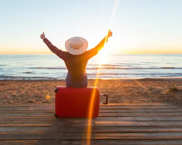 Personne assise sur une valise rouge sur une plage au coucher du soleil, bras levés en signe de victoire.