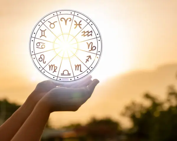 Dłonie trzymają koło zodiaku, gdzie widoczny jest symbol Wagi. Słońce przebija się przez niebo, tworząc magiczną aurę.