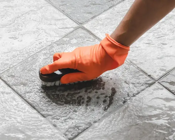 Mano con guanto arancione che spazzola un pavimento piastrellato bagnato, mostrando come togliere ruggine dal pavimento con schiuma.