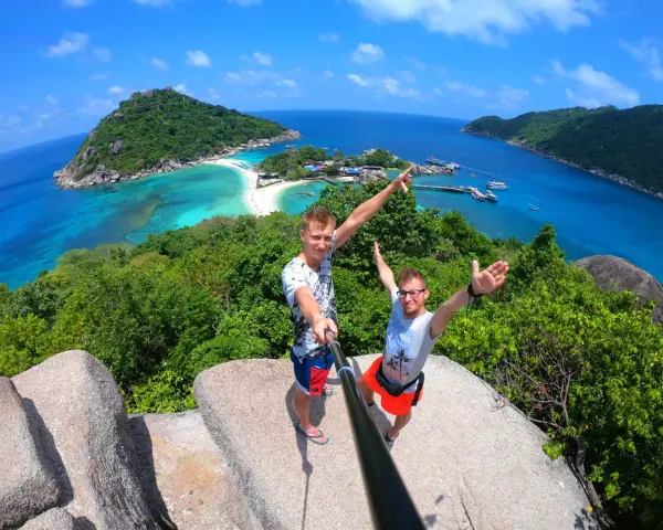 Dwóch mężczyzn robi selfie na tle tropikalnej wyspy z turkusową wodą i plażą.