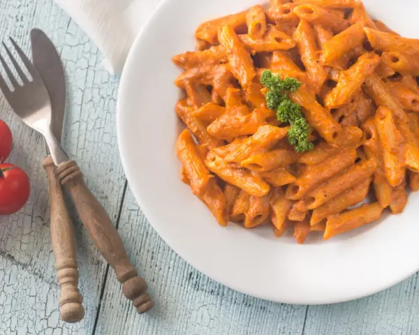 Piatto di penne alla vodka cremose, guarnite con prezzemolo, su un tavolo di legno azzurro. Accanto, pomodorini freschi e posate rustiche.