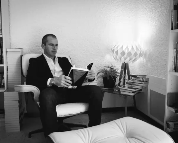 Un homme lit un livre dans un fauteuil, entouré de livres. La lumière d'une lampe éclaire la scène. ae-l af-l.