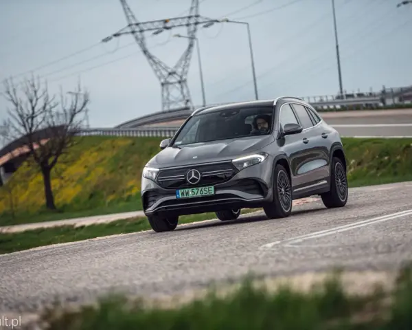 Szary Mercedes-Benz EQA jedzie po drodze, w tle linie energetyczne i drzewo.
