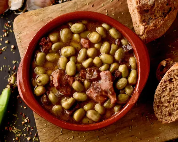 Un cuenco de barro con habas a la catalana, trozos de tocino y pan rústico al lado.