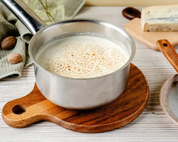 Cremige Gorgonzola-Sauce in einem Topf, bestreut mit Gewürzen. Daneben liegen Muskatnüsse und ein Stück Gorgonzola.