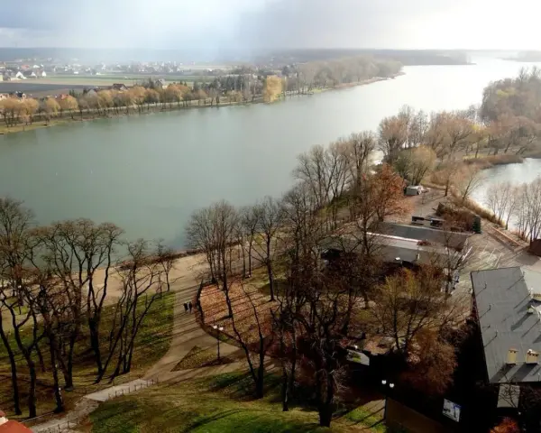 Widok na jezioro nadrowskie z wyspą i zabudowaniami. Drzewa bez liści, jesienny krajobraz.