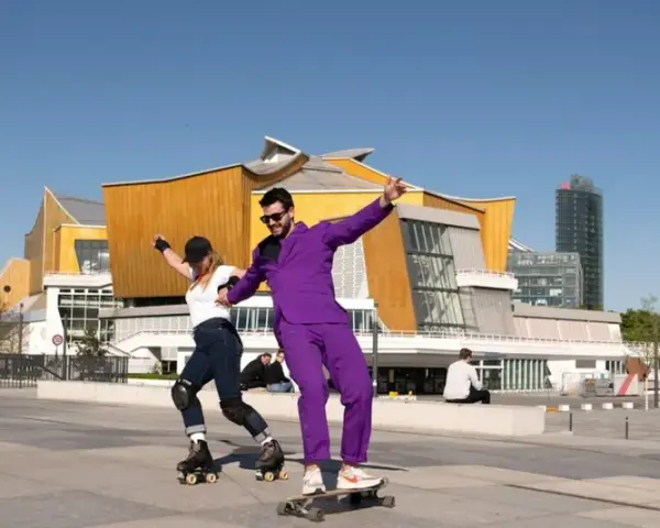 Ein Mann im lila Anzug auf einem Skateboard und eine Frau auf Rollschuhen tanzen vor der Philharmonie. Ein sonniger Tag heute in Berlin.