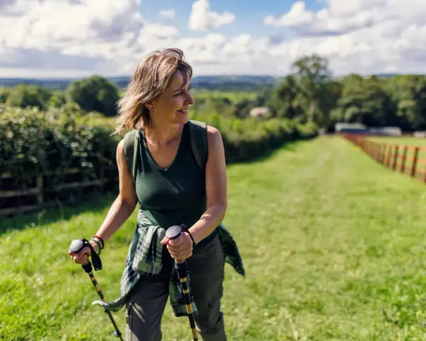 Donna sorridente con bastoncini da nordic walking, che lavora i muscoli coinvolti in un paesaggio verdeggiante.
