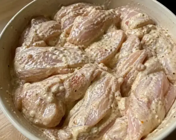 En skål med yoghurtmarinerad kyckling, redo att tillagas. Kycklingbitarna är täckta av en krämig, kryddig marinad.