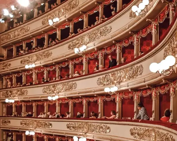 Widownia La Scali, pełna widzów w lożach, oczekuje na rozpoczęcie spektaklu. Bogaty repertuar teatru przyciąga miłośników sztuki.