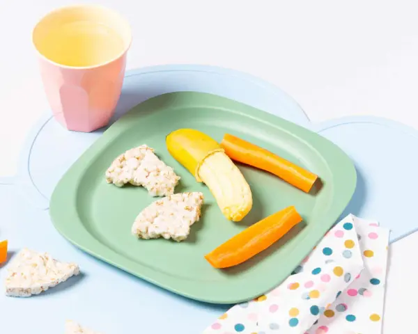 Baby-gerechte Snacks: Reiswaffeln, Karottensticks und eine halbe Banane auf einem grünen Teller.