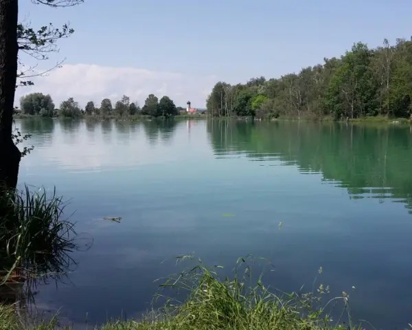 Idyllischer ersinger badesee mit grünen Ufern und einem Kirchturm in der Ferne.