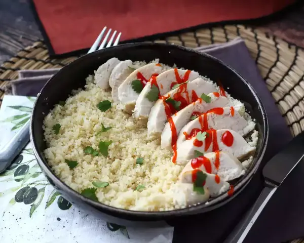 Delizioso petto di pollo in padella, affettato e servito su un letto di couscous, guarnito con prezzemolo e un filo di salsa piccante.