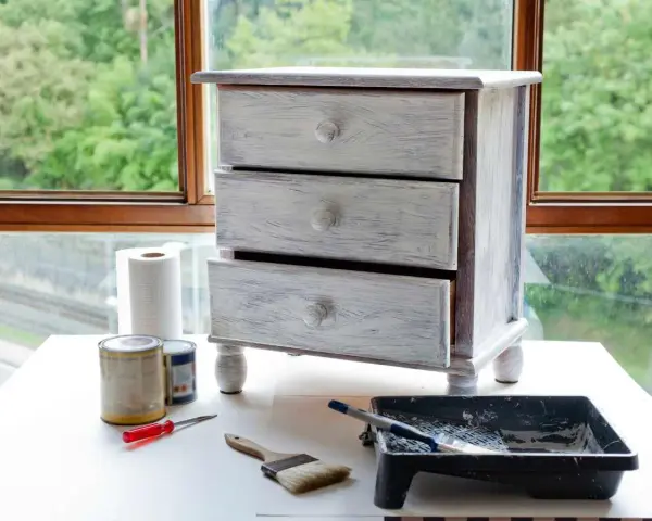 Mueble de tres cajones con acabado desgastado, listo para pintar muebles oscuros. Pinceles, rodillo y latas de pintura a su lado.