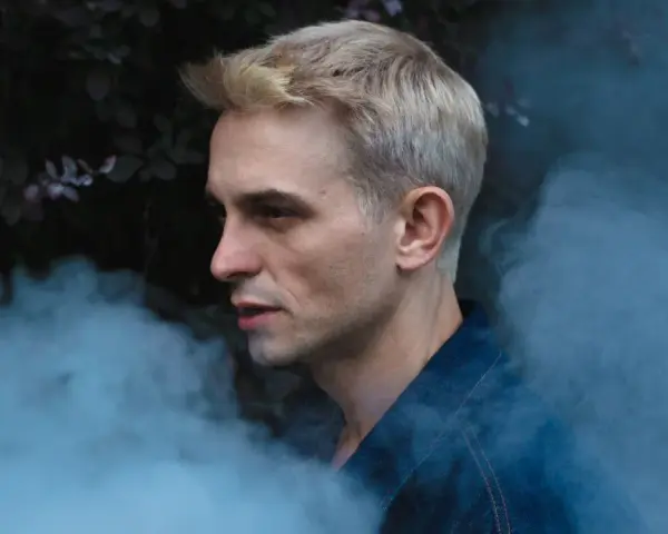 Vito Bambino w dymie, zapowiada nową płytę. Artysta w denimowej kurtce, z blond włosami, na tle ciemnej roślinności.