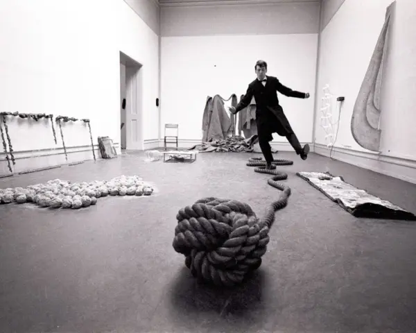 Un homme en costume, peut-être Lawrence Weiner, danse au milieu d'une installation artistique avec des cordes et des objets.