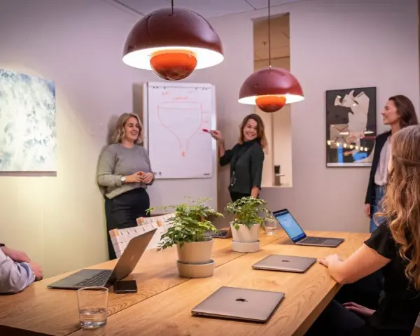Ett team diskuterar strategier vid ett konferensbord med flera laptops. En kvinna pekar på en trattmodell på en whiteboard. Ups datorn är en del av deras effektiva arbetsflöde.