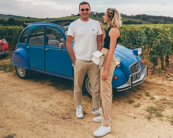 Couple souriant devant une 2CV bleue dans les vignes, une belle idée pour visiter Beaune en 1 jour.