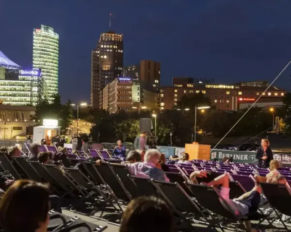 Leute entspannen sich in Liegestühlen beim **Sommerkino am Potsdamer Platz** vor einer leuchtenden Skyline.