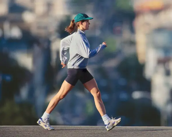 Mujer con gorra y sudadera gris corriendo al aire libre, disfrutando de un paseo después de comer para mantenerse activa.
