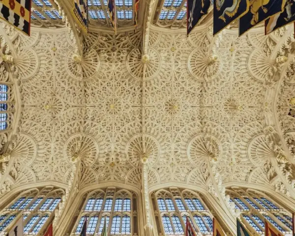 Intricate fan vaulting of a grand hall, reminiscent of the detailed ceilings found in abbey graves, adorned with flags.