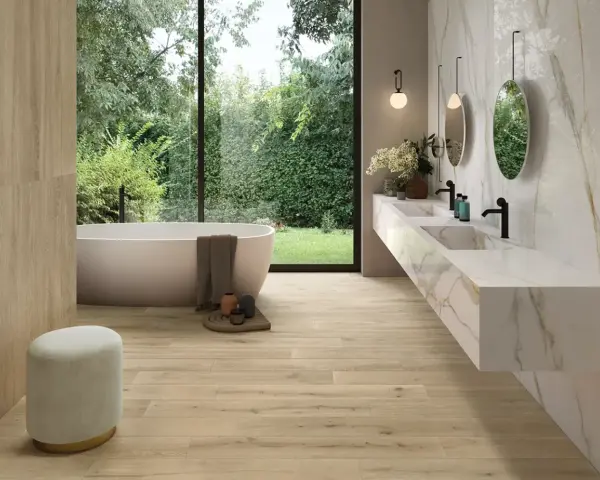 Salle de bain moderne avec baignoire îlot, double vasque en marbre et **pose carrelage imitation parquet** au sol et sur un mur. Vue sur jardin luxuriant.