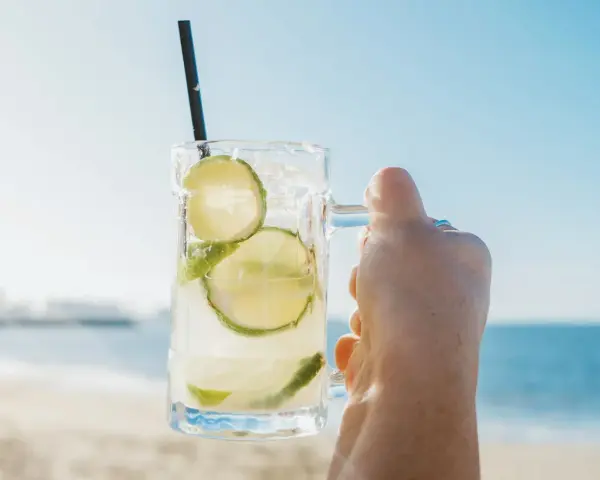 Orzeźwiająca Caipirinha czy Caipiroska z limonką i lodem, trzymana na tle plaży i błękitnego nieba. Idealny napój na wakacje.
