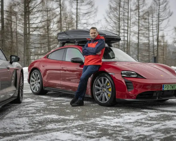 Mężczyzna w czerwonej kurtce opiera się o czerwonego Porsche Taycan z boxem dachowym.