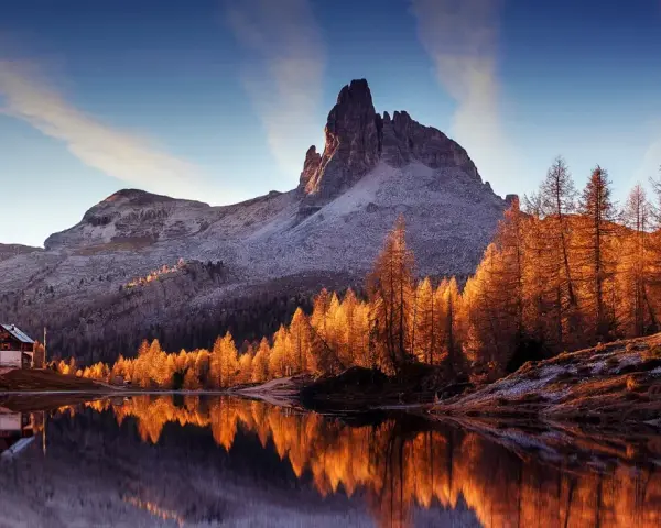 Riflessi dorati sull'acqua di un lago alpino, con un rifugio e montagne innevate. Perfetto per le camminate a Cortina d'Ampezzo.