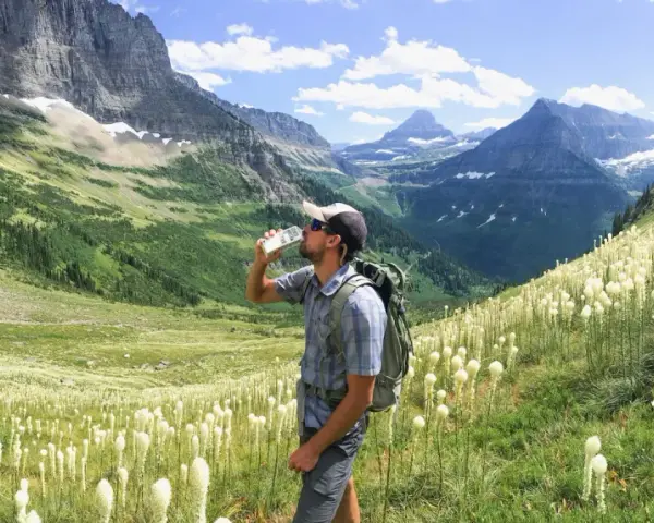 Randonneur buvant de l'eau au milieu d'un champ de fleurs blanches, avec des montagnes majestueuses en arrière-plan. Il espère que cela soulagera sa crampe cuisse.