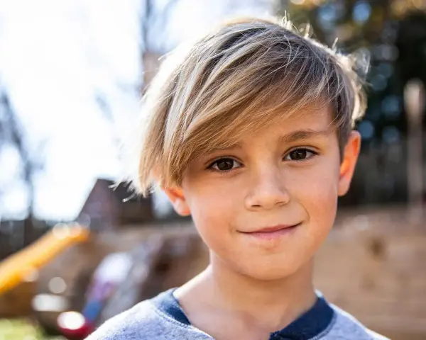 Ein Junge mit blonden Haaren, die nach vorne fallen, lächelt in die Kamera.