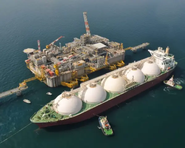 Statek LNG przycumowany do platformy wiertniczej na morzu, holowany przez holowniki.