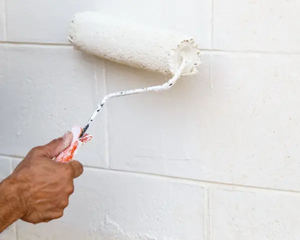 Main tenant, avis peinture carrelage sol : un rouleau blanc applique une nouvelle couche sur un mur en parpaings.