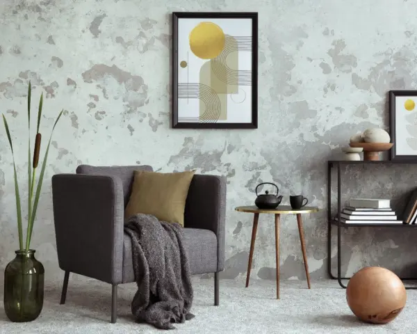 Salón moderno con pared texturizada que combina papel pintado y pintura. Sillón gris, mesa auxiliar dorada, estantería y arte abstracto.
