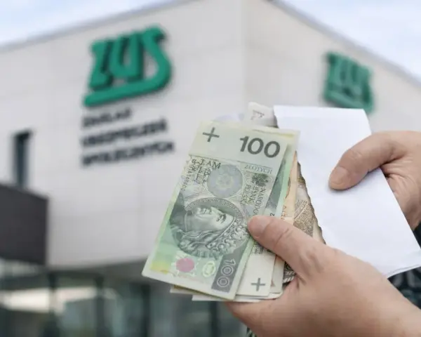 Ręce trzymają polskie banknoty przed budynkiem ZUS. To symboliczny moment, kiedy wypłata 14 emerytury staje się rzeczywistością.