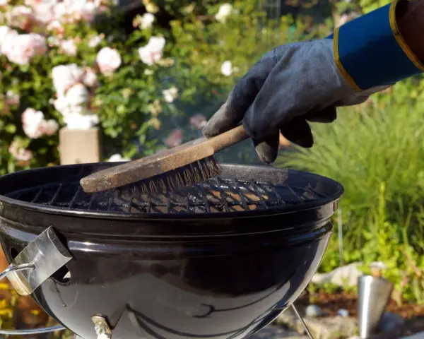 Mit einem Handschuh wird der Grillrost gereinigt. Rauch steigt auf, während die Bürste über die Roste fährt.