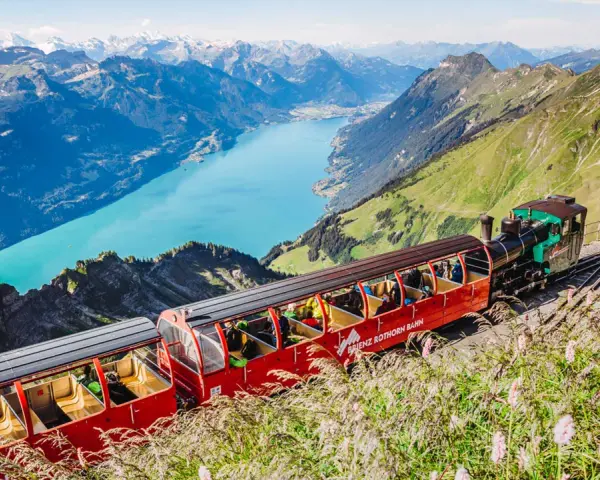 The Brienzer Rothorn train ascends a steep mountain track, offering passengers breathtaking views of a turquoise lake and snow-capped peaks.