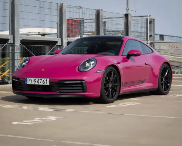 Różowy Porsche 911 Carrera 4S z czarnymi felgami i naklejką PORSCHE na progu, zaparkowany na zewnątrz.