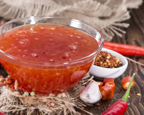Schüssel mit leuchtend roter Sweet Chili Sauce, umgeben von frischen Chilis, Knoblauch und Gewürzen. Perfekt für ein Sweet Chili Sauce Rezept!