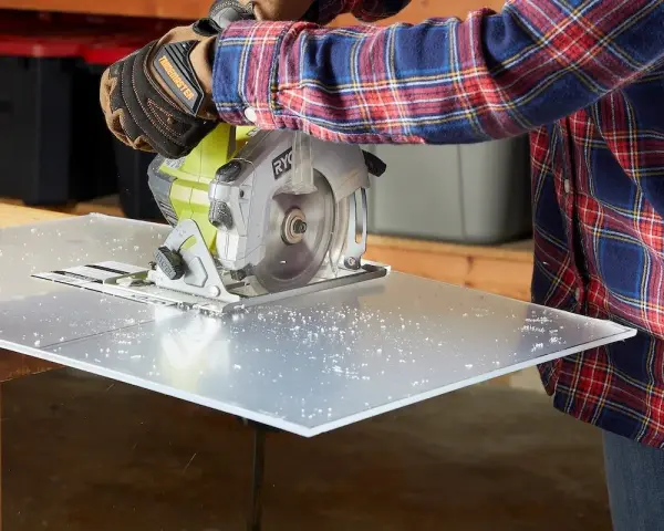 A person in a plaid shirt and gloves uses a circular saw to cut a sheet of plexiglass, creating small plastic shavings. This demonstrates that plexiglass is easy to cut with the right tools.