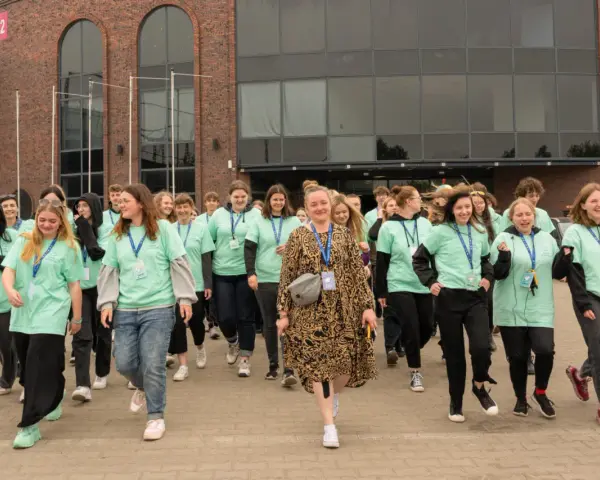 Grupa młodych ludzi w zielonych koszulkach z napisem "Szkoła w chmurze przedmioty" idzie przed budynkiem.